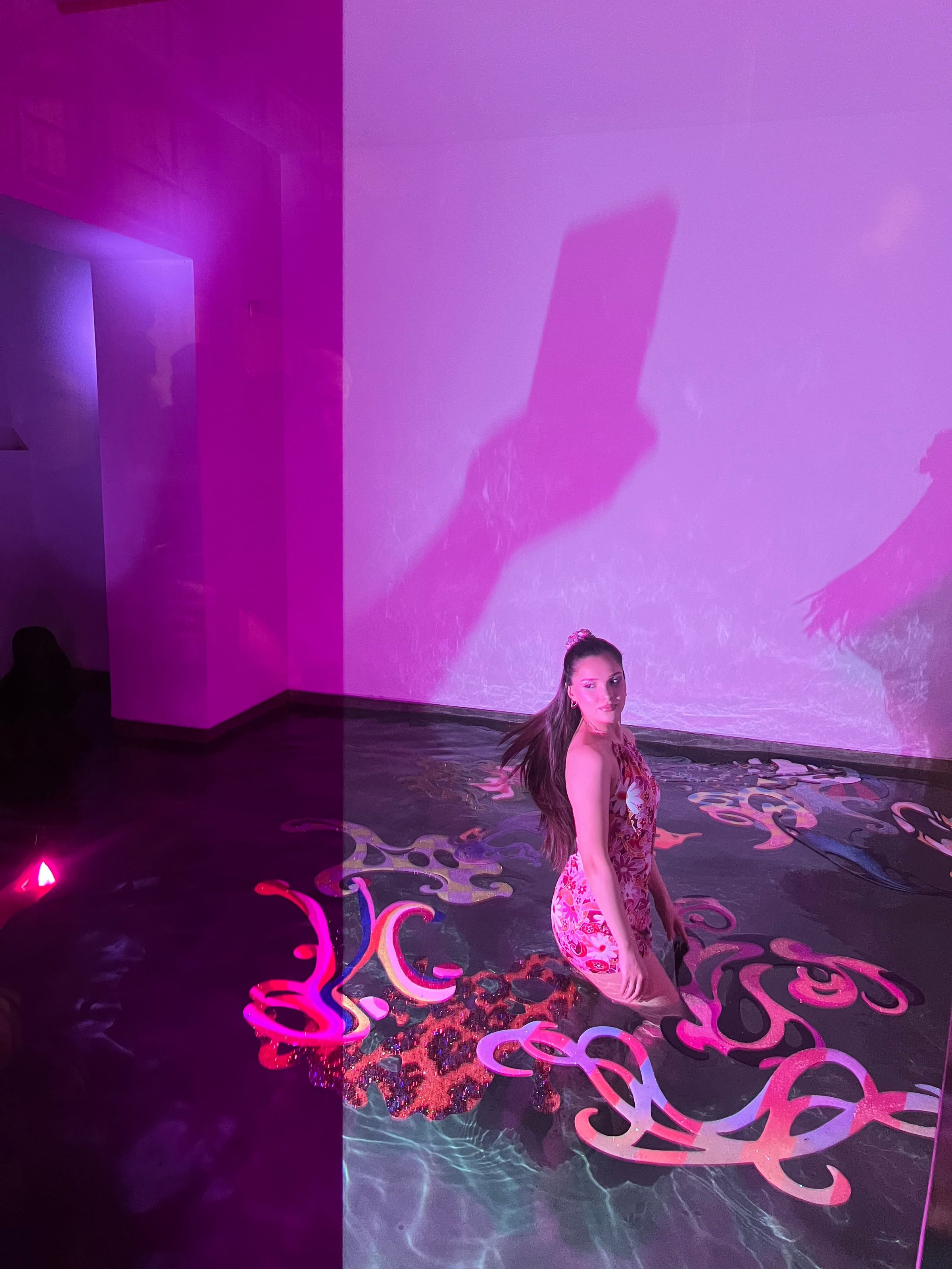 Una persona con vestido estampado posa en el agua entre las piezas, bajo luz magenta